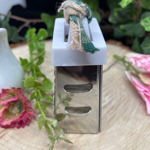 May include: A small, silver grater with a white plastic top and a green and white fabric loop. The grater has two rows of holes and is sitting on a wooden surface.