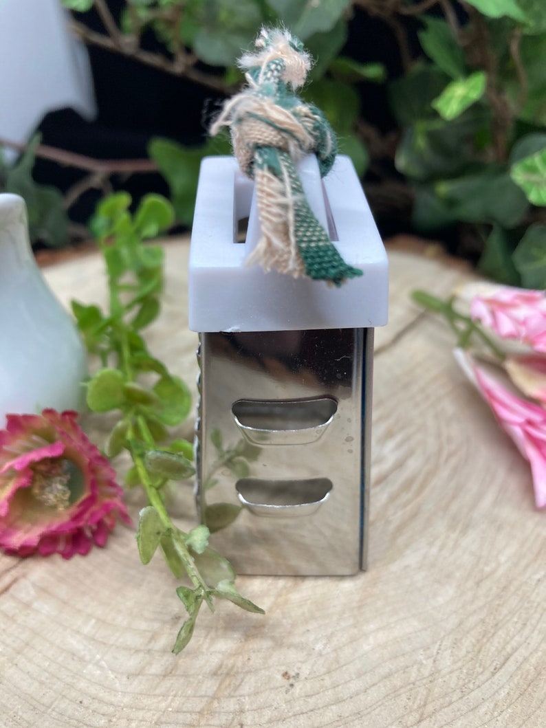 May include: A small, silver grater with a white plastic top and a green and white fabric loop. The grater has two rows of holes and is sitting on a wooden surface.