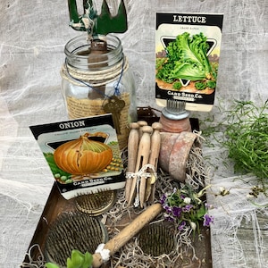 May include: A vintage metal tray with a rustic finish, filled with gardening supplies.  The tray contains a glass jar with a green metal fork, a wooden dowel, a small terracotta pot, and three vintage seed packets for lettuce, onion, and yellow globe dandelions. The seed packets feature colorful illustrations of the vegetables and the text "Card Seed Co."