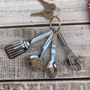 Op de afbeelding: Een zilveren sleutelhanger met een set miniatuur keukengerei, waaronder een spatel, een geperforeerde lepel, een lepel en een garde.