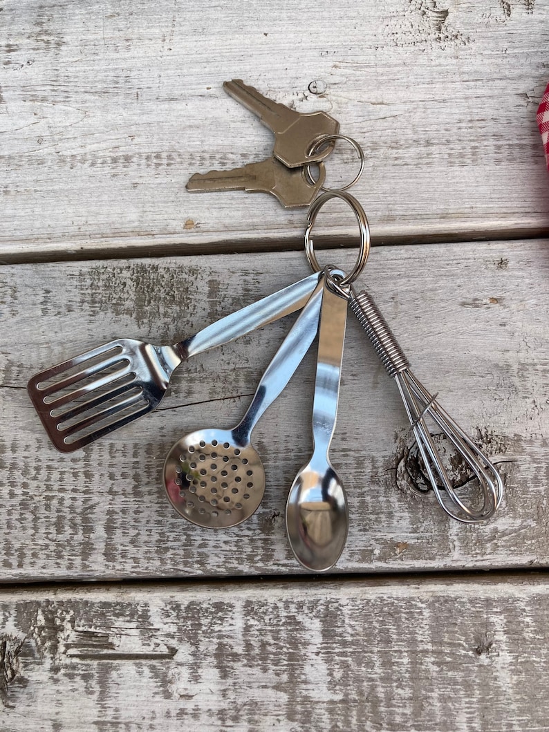 Op de afbeelding: Een zilveren sleutelhanger met een set miniatuur keukengerei, waaronder een spatel, een geperforeerde lepel, een lepel en een garde.