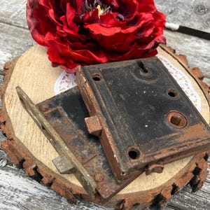 May include: Two rectangular metal rim locks with a weathered black and brown finish. Each lock features a central square spindle hole, mounting screw holes, and a protruding latch mechanism. The metal surfaces show signs of oxidation and age.