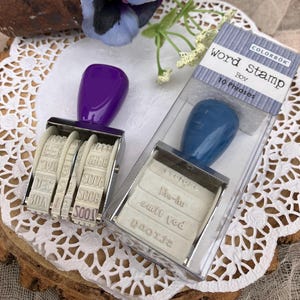 May include: Two rubber stamps, one purple and one blue, with a clear plastic case. The purple stamp has four wheels with numbers and letters. The blue stamp has four lines of text that read "The-du", "omit vod", "2f2002", and "Σ". The case has the text "COLORBOK word stamp Boy 10 Phrases".