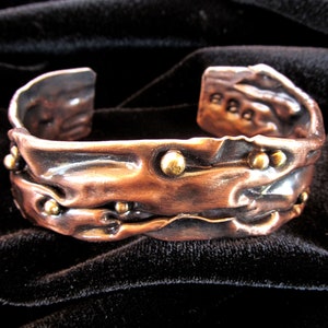 May include: A hammered copper bracelet with a textured surface and gold-toned accents. The bracelet is open and has a wide, flat band.