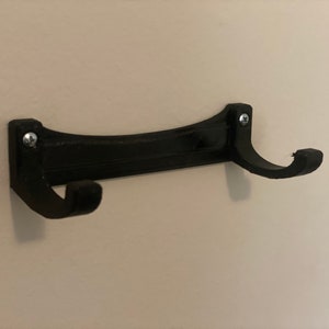 May include: A black wall-mounted coat rack with two curved hooks. The rack is made of a solid material and is attached to the wall with two screws. The design is simple and functional, suitable for hanging coats, hats, or other items.