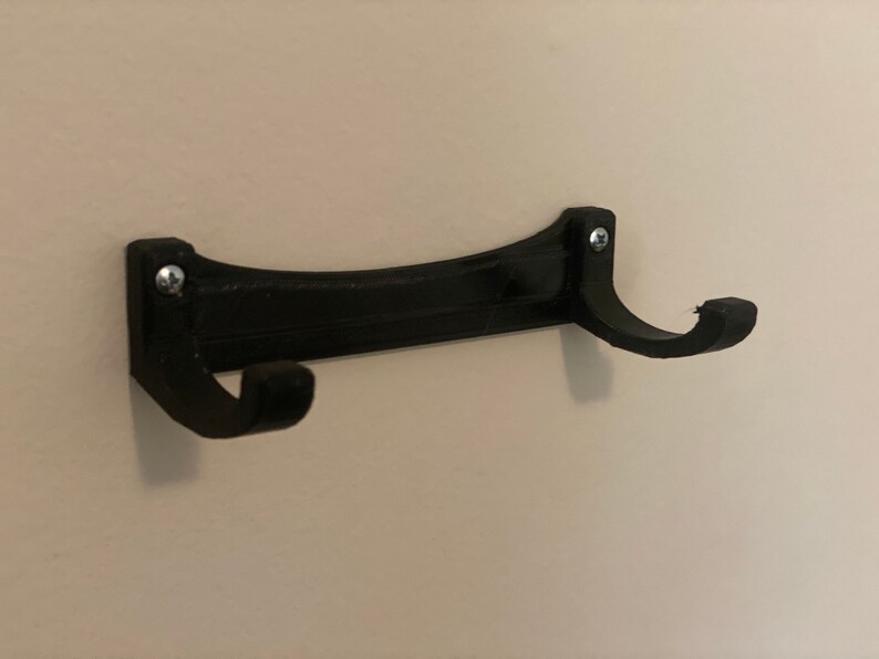 May include: A black wall-mounted coat rack with two curved hooks. The rack is made of a solid material and is attached to the wall with two screws. The design is simple and functional, suitable for hanging coats, hats, or other items.