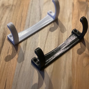 May include: Two 3D-printed wall hooks. One is white, the other is black. Each hook has a curved top and a flat base with two holes for mounting. The hooks are made of plastic and sit on a wooden surface.