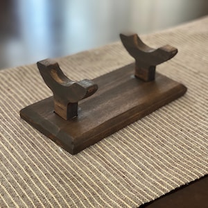 May include: A wooden stand with two curved arms for holding swords or other long objects. The stand is dark brown and has a smooth finish.