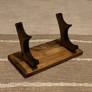 May include: A wooden stand with two curved supports for holding a sword or other long object. The stand is stained a dark brown color.