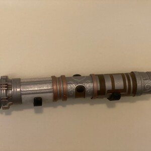 May include: A silver and bronze lightsaber prop with intricate detailing. The hilt features a combination of silver, bronze, and black accents. The lightsaber is mounted on a white wall with two black holders.