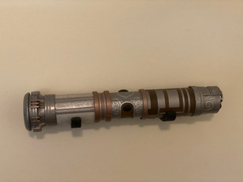 May include: A silver and bronze lightsaber prop with intricate detailing. The hilt features a combination of silver, bronze, and black accents. The lightsaber is mounted on a white wall with two black holders.