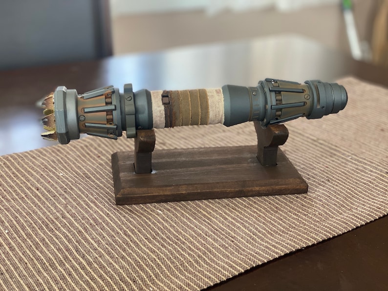 May include: A detailed, gray and brown lightsaber prop with a weathered, fabric-wrapped handle, displayed on a wooden stand.