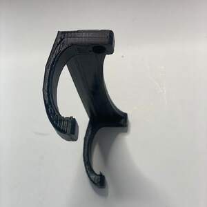 May include: A black, 3D-printed object with a curved design, featuring two hook-like ends and a rectangular top with a small hole. The object appears to be made of plastic and is set against a white background.
