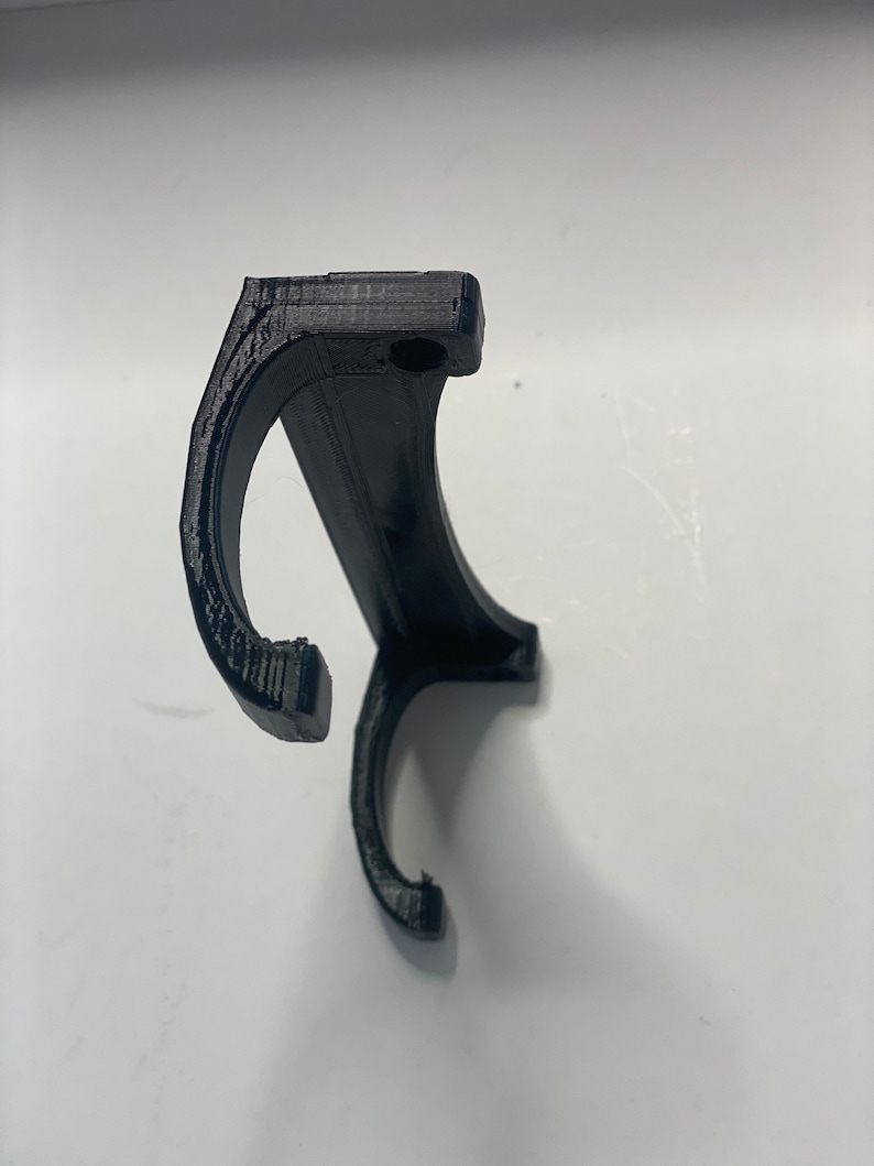 May include: A black, 3D-printed object with a curved design, featuring two hook-like ends and a rectangular top with a small hole. The object appears to be made of plastic and is set against a white background.