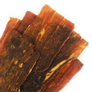 May include: Close-up of several thin, dark brown, dried meat strips stacked on top of each other. The meat is slightly curled and has a rough texture.