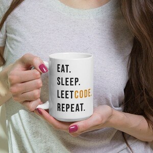 May include: White ceramic mug with the text "EAT. SLEEP. LEETCODE. REPEAT." in black and gold lettering. The mug is held by a person with long brown hair.