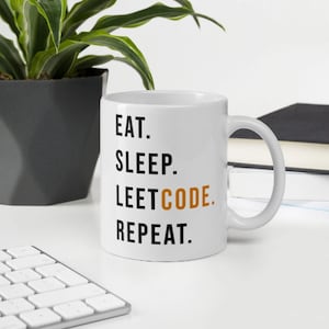 May include: White ceramic mug with the text "EAT. SLEEP. LEETCODE. REPEAT." in black and orange lettering. The mug is on a white surface with a keyboard, a potted plant, and a stack of books in the background.