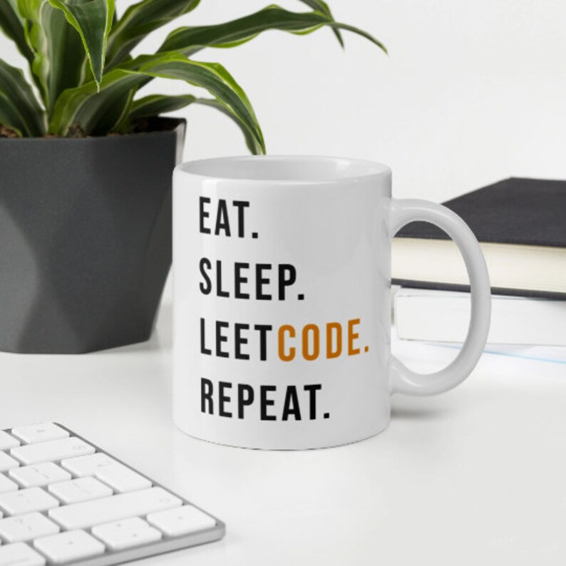 May include: White ceramic mug with the text "EAT. SLEEP. LEETCODE. REPEAT." in black and orange lettering. The mug is on a white surface with a keyboard, a potted plant, and a stack of books in the background.