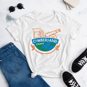 May include: White t-shirt with a graphic design featuring a stylized image of a beer mug, cannon, and a globe with the text "CUMBERLAND COUNTY".