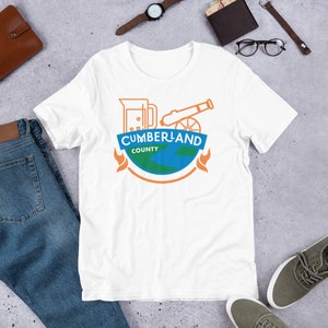 May include: White t-shirt with a graphic design of a beer mug, cannon, and globe with the text "CUMBERLAND COUNTY".
