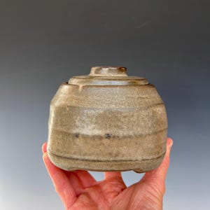 May include: A small, round, ceramic jar with a lid. The jar is a light brown color with a speckled glaze. The lid is slightly darker brown.
