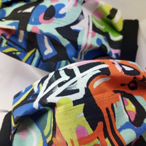 Op de afbeelding: Twee kleurrijke gezichtsmaskers met abstracte patronen in tinten blauw, groen, geel, oranje, roze en zwart. De maskers hebben zwarte elastische oorlussen.