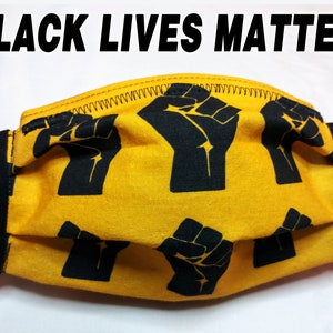 May include: A yellow face mask with a black fist pattern and black trim. The text "BLACK LIVES MATTER" is printed in bold black letters above the mask. The mask has white elastic ear loops.