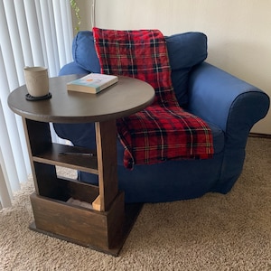 May include: A dark brown wooden end table with a round top and a shelf underneath. The table has a storage compartment with a wooden door. A blue armchair with a red and black plaid throw blanket is next to the table.