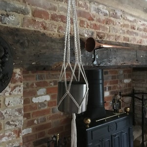 Puede incluir: Un macetero de macramé blanco con una maceta de metal colgando de él. El macetero está colgado de una viga de madera en una habitación de ladrillo.