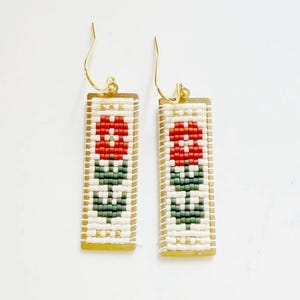 May include: A pair of rectangular beaded earrings with a floral design. The earrings feature a red flower, green leaves, and white background, all set against a gold-toned frame. The earrings hang from gold-colored ear wires.