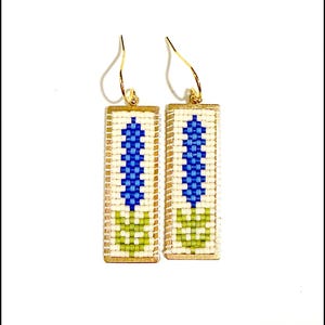 May include: A pair of rectangular earrings with gold-colored hooks. Each earring features a cream-colored background with a blue and green floral design. The earrings are made with small beads.