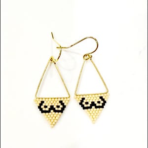 May include: A pair of gold-toned triangle earrings with a beaded design. The bottom of each earring features a cream-colored beaded pattern with a black geometric design. The earrings have a hook closure.