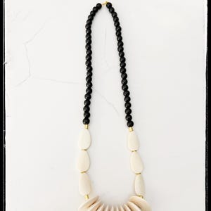 May include: A necklace featuring a strand of small, black beads connected to cream-colored, oval-shaped beads. The necklace has a gold-colored accent at the connection points and a fan-shaped pendant made of cream-colored, flat, pointed pieces.