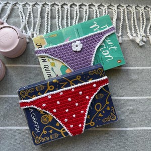 Puede incluir: Dos braguitas de bikini de ganchillo. Una es de color morado claro con un detalle de flor blanca de ganchillo. La otra es roja con lunares blancos y un borde blanco de ganchillo. Ambos artículos están sobre dos libros con texto e ilustraciones.