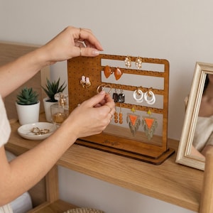 Peut inclure: Présentoir à boucles d'oreilles en bois marron avec des trous et des fentes horizontales. Le présentoir porte le nom "Jennifer". Plusieurs paires de boucles d'oreilles de différents styles et couleurs sont accrochées.