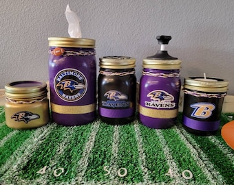 May include: Set of five purple and gold mason jars decorated with the Baltimore Ravens logo. The jars are used for various purposes, including a tissue holder, a soap dispenser, a candle holder, and two other jars.