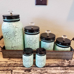 May include: Set of five turquoise blue Ball mason jars with black lids and crystal knobs. The jars are arranged in a metal tray with a wooden handle. The jars are labeled "Ball Wide Mouth" and "Ball Mason".