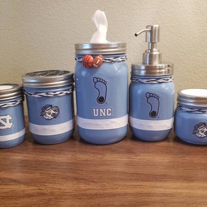 May include: Set of five blue glass jars with white and black accents. The jars are decorated with UNC logos and a basketball. The jars are arranged in a row on a wooden surface.