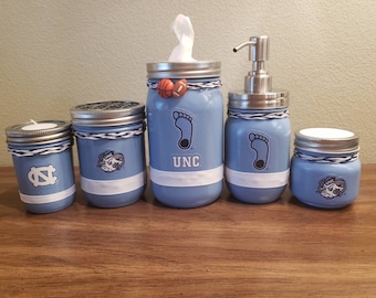 May include: Set of five blue glass jars with white and black accents. The jars are decorated with UNC logos and a basketball. The jars are arranged in a row on a wooden surface.