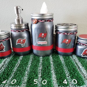 May include: Set of four gray glass jars with red and white accents and a Tampa Bay Buccaneers logo. The jars are decorated with a pirate ship and the words "Buccaneers".