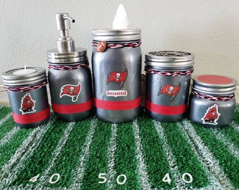 May include: Set of four grey glass jars with red and white accents and a Tampa Bay Buccaneers logo. The jars are decorated with a pirate ship and the words "Buccaneers".
