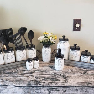 May include: White painted mason jars with black lids and black ribbon accents. The jars are arranged on a galvanized metal tray with wooden handles. The jars are labeled with the word "Ball" and some have additional labels like "Bake" and "Spice".  There are also black and silver kitchen utensils in the jars.