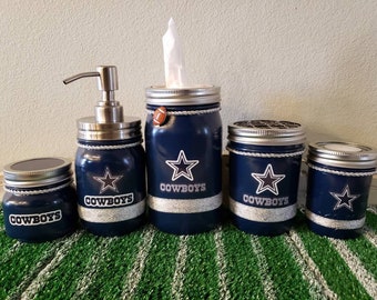 Op de afbeelding: Set van vijf blauwe glazen potten met zilveren deksels en een zilveren pompflacon. Elke pot heeft een zilveren ster en het woord "COWBOYS" erop geschreven. De potten staan op een groen-wit gestreept oppervlak.