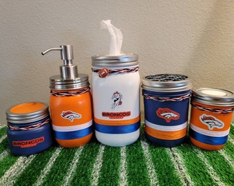 May include: Set of five bathroom jars decorated with the Denver Broncos logo and team colours. The jars include a soap dispenser, a tissue holder, a toothbrush holder, a cotton swab holder, and a candle holder.