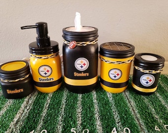 May include: Set of four black and yellow mason jars with the Pittsburgh Steelers logo and the word "Steelers" on each jar. The jars are decorated with black and yellow stripes and twine. The jars are on a green and yellow surface.