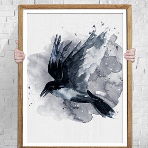 May include: A framed watercolor print of a crow in flight. The artwork features a black and gray color palette with a splash effect. The crow is depicted with its wings spread, set against a backdrop of gray watercolor washes. The frame is a light brown color.