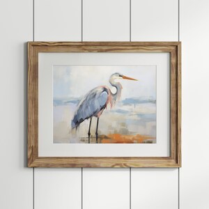 Puede incluir: Una pintura enmarcada de una garza azul de pie en una playa con un fondo borroso de agua y cielo.