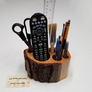 May include: A wooden desk organizer shaped like a tree trunk with multiple compartments. The organizer holds a remote control, scissors, pens, pencils, and a paintbrush.