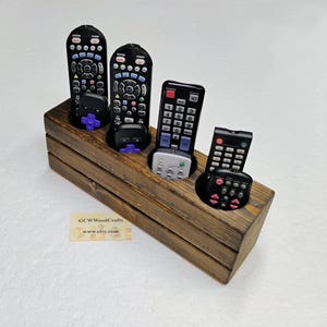 May include: A rustic wooden remote control holder with four circular openings, each holding a black remote. The holder is made of stained wood and has a rectangular shape. The text "GCWWoodCrafts" and "www.etsy.com" is visible on a small label.
