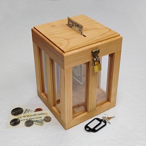 May include: A wooden money bank with a cage-like design. The bank has a slot on top for inserting bills and a small lockable door. A dollar bill is visible in the slot. The bank is made of light-colored wood.